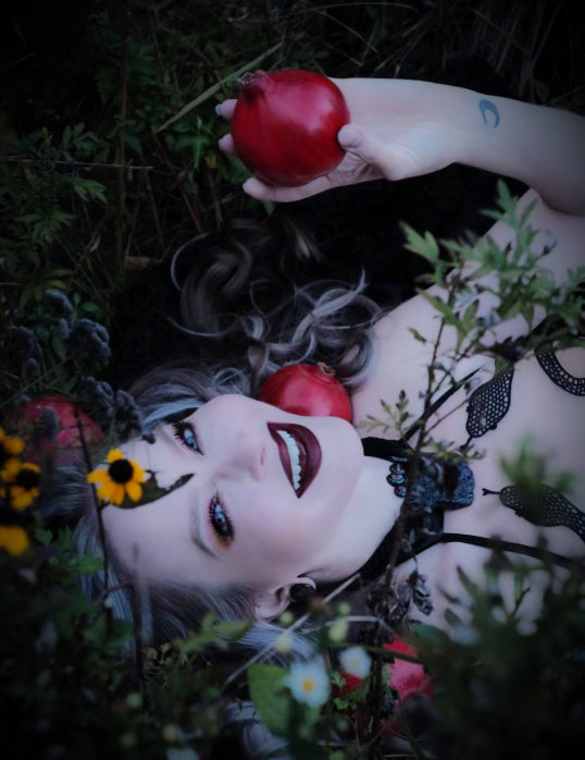 A blond woman with blue eyes and dark makeup is laying in a dark field of yellow flowers and holding a bright red pomegranate.  She is wearing a bodysuit with snakes crawling up it and a black gemstone necklace and is also surrounded by pomegranates.  